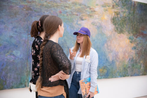 Auf dem Foto sind drei Frauen zu sehen, sie stehen vor einem grossen, farbigen Gemälde in der Fondation Beyeler. Eine der drei Frauen erzählt den beiden anderen gerade etwas und gestikuliert dabei mit ihrer rechten Hand.