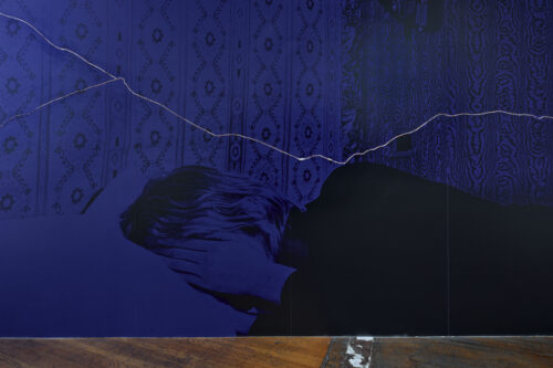 Das Bild zeigt ein blau eingefärbte Fotografie von einer weiblich gelesenen Person, die im Bett liegt und sich die rechte Hand vor das Gesicht hält. Im Hintergrund ist eine Wandfassade mit komplexen Mustern zu sehen. Zudem verläuft auf der Fotografie von links nach rechts eine schmale weisse Linie.