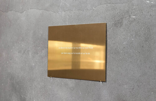 Das Bild zeigt eine graue Betonwand, auf der ein rechteckiges goldenes Schild angebracht ist. Darauf steht geschrieben: Le devoir de respirer sur ma tète sur mon front sur mon sang sur le monde et sur la vie.