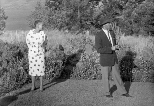 Eine historische Schwarz-Weiss-Fotografie zeigt eine Frau und einen Mann, die auf einer Grasfläche stehen. Im Hintergrund ist ein üppiger Wald zu sehen. Beide Personen blicken nach links, die Frau trägt ein modisches zweiteiliges Kleid mit grafischem Muster, der Mann einen Anzug, Krawatte und Hut.
