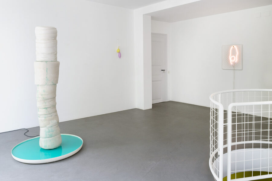 In einem weissen Raum sind drei Kunstwerke zu sehen. Links eine säulenartige Struktur, in der Mitte zwei Luftballons und rechts eine Neonarbeit.