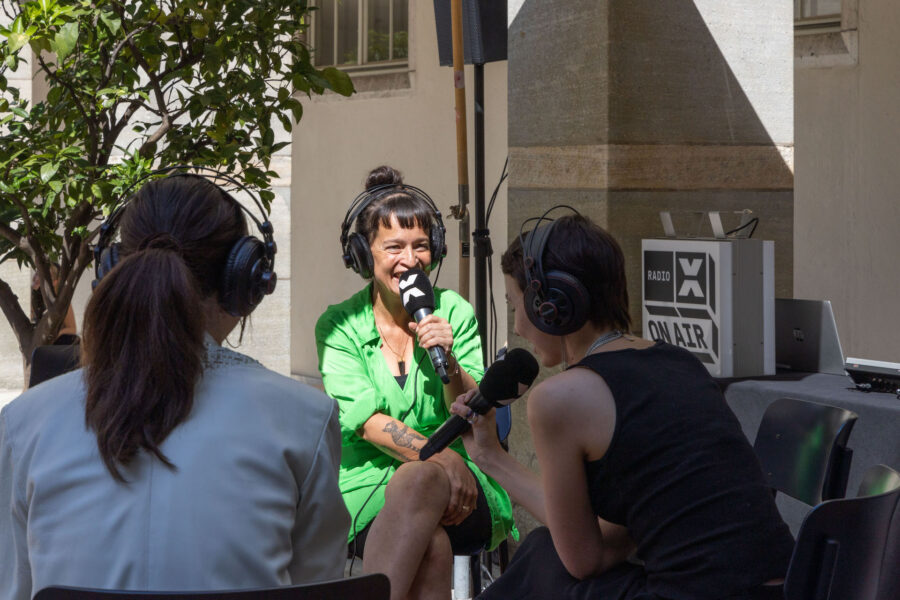 Das Bild zeigt eine Interviewsituation unter freiem Himmel bei Sonnenschein. Zu sehen sind drei Frauen, sie tragen einen Kopfhörer und halten ein Mikrofon in der Hand.