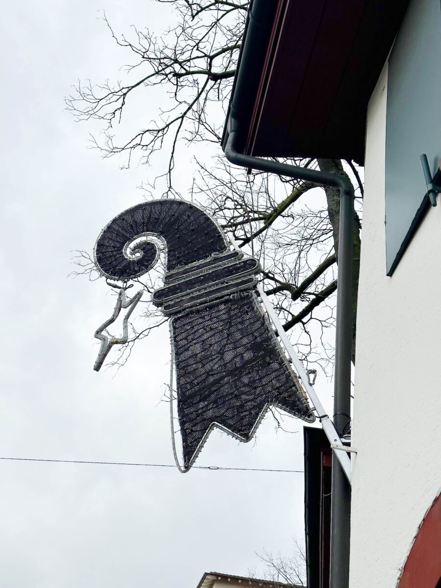 Das Bild zeigt eine Hausfassade, an der ein Fahnenständer angebracht ist. Am Fahnenständer hängt ein grosser Baslerstab, umgeben von einer ausgeschalteten Lichterkette.