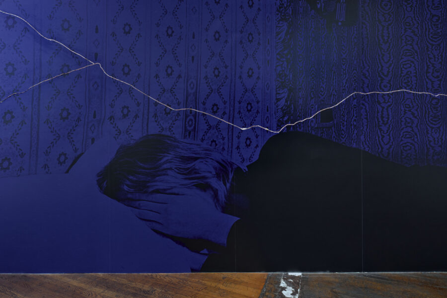 Das Bild zeigt ein blau eingefärbte Fotografie von einer weiblich gelesenen Person, die im Bett liegt und sich die rechte Hand vor das Gesicht hält. Im Hintergrund ist eine Wandfassade mit komplexen Mustern zu sehen. Zudem verläuft auf der Fotografie von links nach rechts eine schmale weisse Linie.