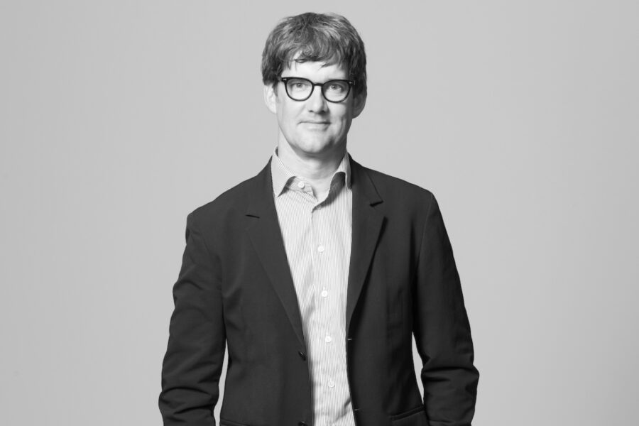 Die Schwarz-Weiss-Fotografie zeigt einen Mann im mittleren Lebensalter mit freundlichem, geschlossenem Lächeln, schwarzer Brille und kurzem Haar. Er trägt ein gestreiftes Hemd und einen schwarzen Anzug.
