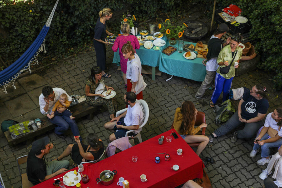 Das Bild zeigt eine Menschengruppe in ausgelassener Stimmung in einem Hintergarten. Auf einem Tisch sind mehrere Mahlzeiten auf Tellern zu sehen, daneben auch ein Grill. An einem anderen Tisch und auf Holzbänken sitzen Personen, die essen und miteinander sprechen.