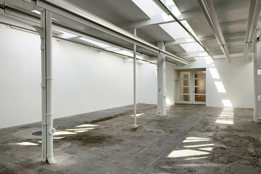 Das Bild zeigt einen leeren Raum mit Steinboden, mehreren stehenden Balken und einer Türe im Hintergrund. Die Wand ist weiss gestrichen, ein Teil der Decke ist mit Fenstern ausgestattet, der Boden reflektiert an gewissen Stellen das Sonnenlicht.