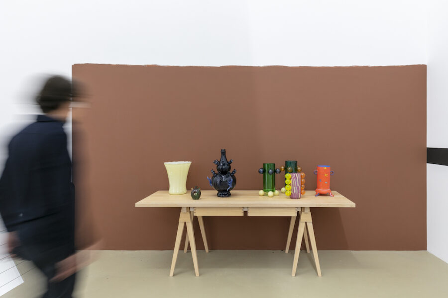 Das Bild zeigt einen braunen Holztisch mit verschiedenen bunten Vasen darauf, einige davon sind aus Glas. Der Tisch befindet sich vor einer braun bemalten Wandfläche. Links im Vordergrund ist eine verschwommene Person in schwarzer Kleidung zu sehen, die gerade auf den Tisch zugeht.