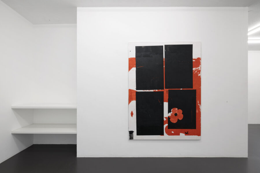 Das Bild zeigt eine weisse Wand in einer Galerie. An der Wand hängt ein Gemälde, darauf sind vier schwarze Rechtecke zu sehen, umgeben von weisser und hellroter Farbe. In einem der vier Rechteck ist eine Blume abgebildet, ebenfalls in den Farben weiss und hellrot.