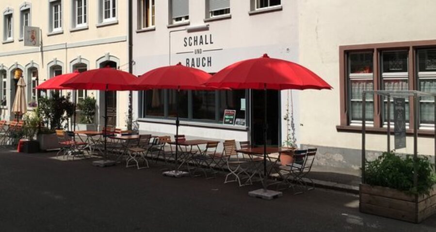 Das Bild zeigt den Vordereingang eines Restaurants. Die Sonne scheint. Vor dem Betrieb sind Stühle, Tische und grosse rote Sonnenschirme zu sehen.